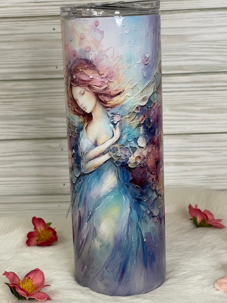 May include: A stainless steel tumbler featuring a watercolor illustration of a woman with flowing hair and a flowing gown. The design incorporates soft blues, purples, and pinks. The tumbler has a clear lid and is suitable for hot or cold beverages.