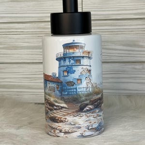 May include: White ceramic soap dispenser with a black pump. The dispenser features a watercolor illustration of a blue lighthouse with a red-tiled roof, set against a coastal scene with seashells.