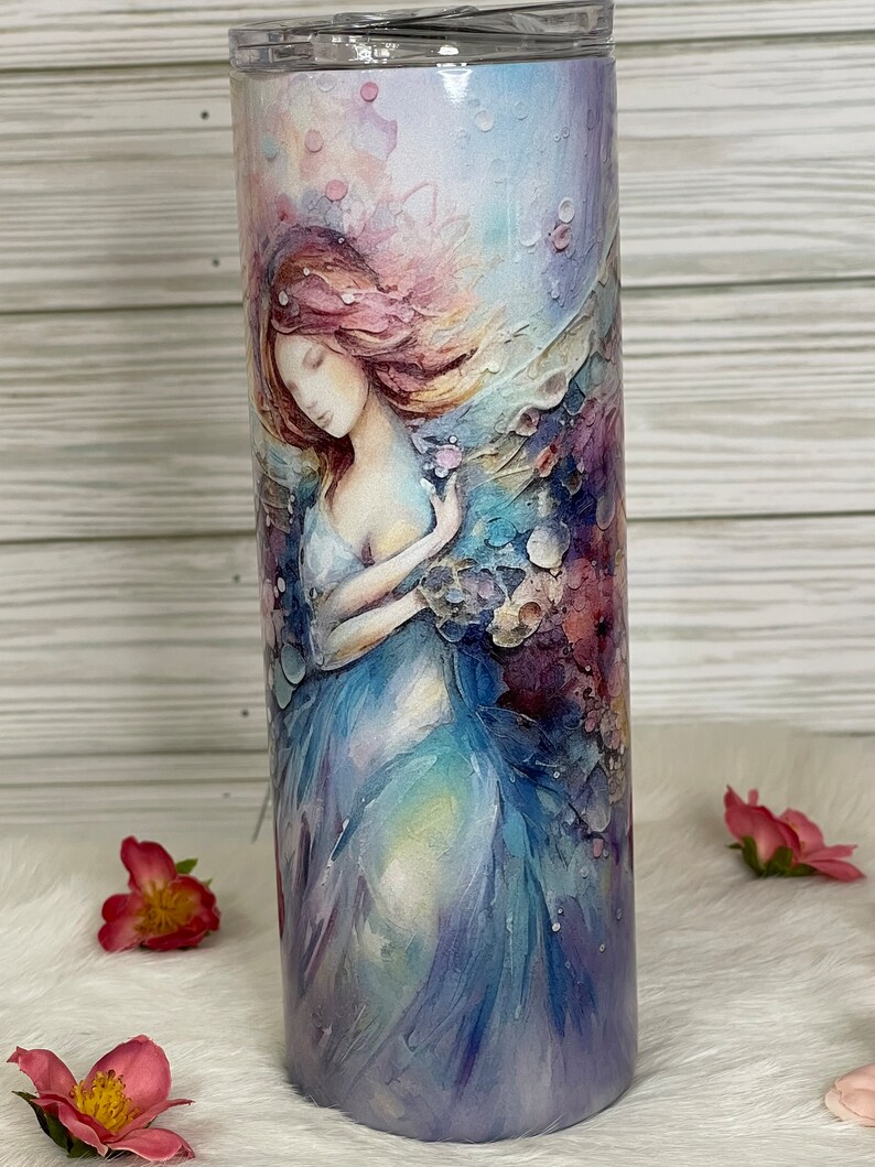 May include: A stainless steel tumbler featuring a watercolor painting of a woman in a flowing blue dress. The artwork has soft pastel colors, including pink, purple, and blue. The tumbler is cylindrical and has a clear lid.