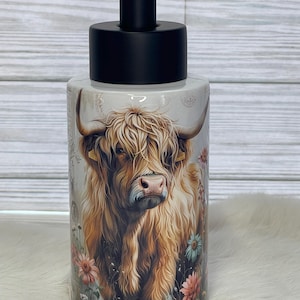 May include: A ceramic soap dispenser with a black pump top. The dispenser features a detailed illustration of a Highland cow with long, shaggy brown fur, surrounded by colorful flowers. The background is a light, neutral color.