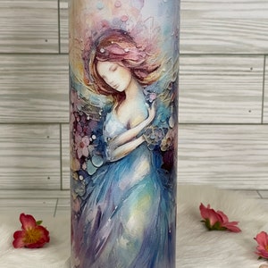 May include: A stainless steel tumbler featuring a watercolor illustration of a woman in a flowing blue dress, surrounded by flowers. The design incorporates soft pastel colors, including pink, purple, and blue, creating a dreamy aesthetic. The tumbler has a clear lid.