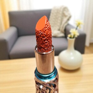 Puede incluir: Primer plano de una barra de labios de color rojo anaranjado. La barra de labios tiene un dise&ntilde;o texturizado y detallado. El tubo met&aacute;lico tiene un dise&ntilde;o de filigrana decorativo y una banda turquesa.
