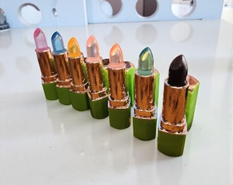 May include: A collection of lipsticks with various colors, including pink, blue, orange, and green. The lipsticks have gold-colored tubes and green bases. The lipstick colors range from light to dark, with one being a deep brown.