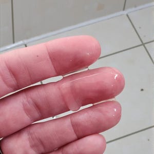 May include: Close-up of a hand with clear, viscous liquid on the fingers. The skin is pinkish-beige, and the liquid glistens, reflecting light. The background is a tiled surface.