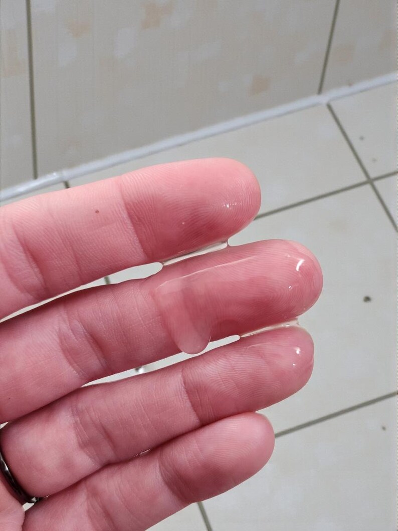 May include: Close-up of a hand with clear, viscous liquid on the fingers. The skin is pinkish-beige, and the liquid glistens, reflecting light. The background is a tiled surface.
