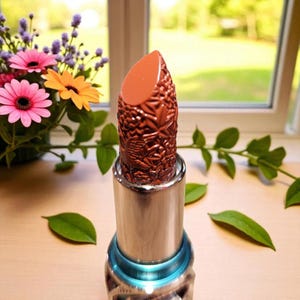Puede incluir: Un labial con un dise&ntilde;o floral marr&oacute;n detallado en el propio labial. El labial est&aacute; en un tubo plateado con un acento azul. El fondo incluye flores y vegetaci&oacute;n.