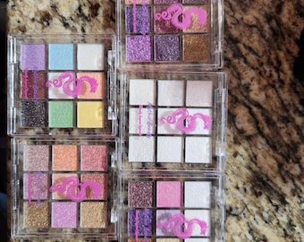 May include: Multiple eyeshadow palettes in clear plastic cases. Each palette contains nine squares of various colors, including pinks, purples, and golds. The text "Mythic Beauty" and a pink dragon logo are printed on each case.