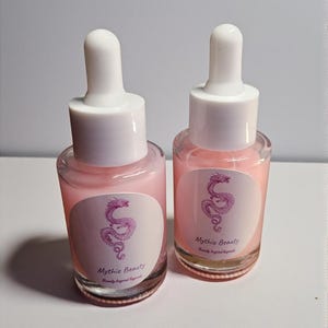 May include: Two glass dropper bottles filled with a pink liquid. Each bottle has a white cap and a label with a pink dragon illustration and the text "Mythic Beauty". The bottles are on a light background.