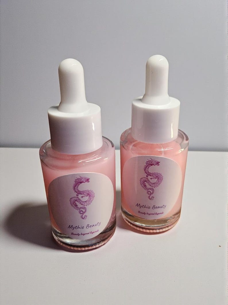 May include: Two glass dropper bottles filled with a pink liquid. Each bottle has a white cap and a label with a pink dragon illustration and the text "Mythic Beauty". The bottles are on a light background.