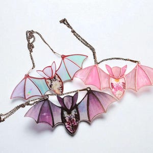 May include: Three bat-shaped necklaces with iridescent wings and floral centers. The wings are in shades of pink, purple, and blue, attached to delicate chains. The bats have a gothic, whimsical aesthetic.
