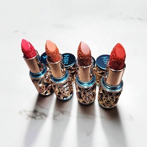 May include: An assortment of lipsticks in various shades of red, coral, and pink. The lipsticks have intricate gold-colored casings with a blue accent. The lipstick tops are textured, and the caps have the letters "HIH" printed on them.
