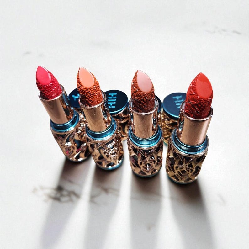Puede incluir: Una variedad de labiales en tonos rojo, coral y rosa. Los labiales tienen estuches dorados intrincados con un acento azul. Las puntas de los labiales son texturizadas y las tapas tienen las letras "HIH" impresas.