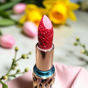 Puede incluir: Un labial rojo con un dise&ntilde;o floral en el labial. El labial est&aacute; en un tubo dorado con una banda azul. El fondo incluye narcisos amarillos y tulipanes rosas.
