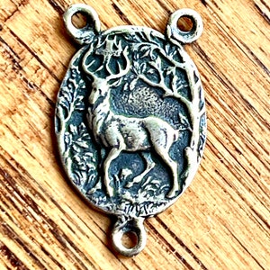 May include: A silver-toned metal charm featuring a detailed embossed image of a deer standing in a forest setting. The charm has two loops for attaching to a necklace or bracelet.