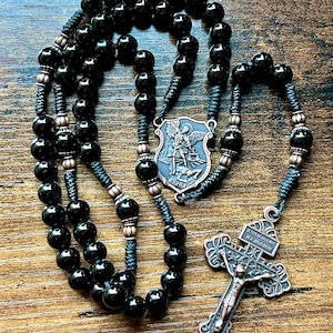 May include: A black beaded rosary with a bronze-colored crucifix and a shield-shaped centerpiece depicting a saint. The beads are strung on black cord, and the crucifix features the figure of Jesus. The rosary is displayed on a wooden surface.