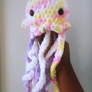 May include: A crocheted jellyfish toy with a pink, yellow, and white body and purple tentacles. The jellyfish has black eyes.