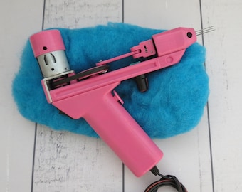 May include: A pink electric felting tool with a blue piece of wool underneath. The tool has a black cord and a small black switch.
