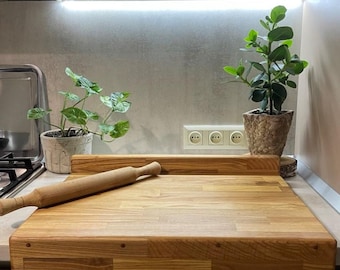 May include: A large wooden cutting board with a rolling pin on top. The board is made of light wood and has a natural finish. The board is sitting on a countertop in a kitchen.