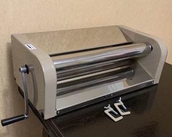 Op de afbeelding: Een beige handmatige pastaroller met twee roestvrijstalen rollen. De roller heeft een handkrans en staat op een donkerbruin oppervlak. Twee metalen klemmen zijn zichtbaar op de voorgrond.