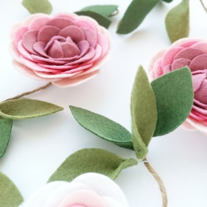 May include: A garland of felt flowers with pink and white petals and green leaves. The flowers are strung together with twine.