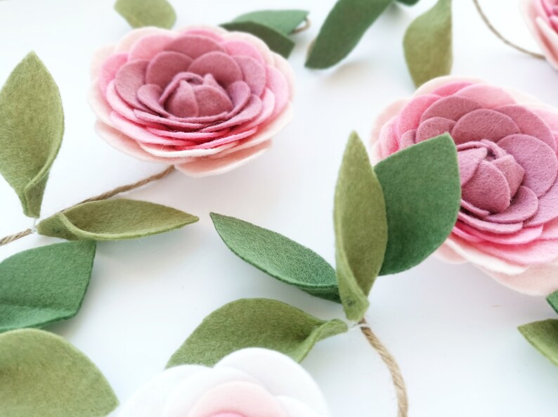 May include: A garland of felt flowers with pink and white petals and green leaves. The flowers are strung together with twine.