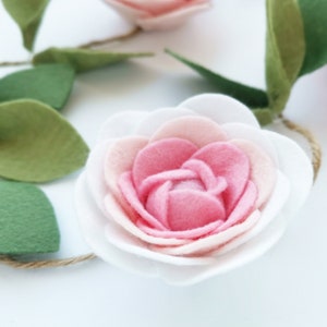 May include: A pink and white felt flower garland with green felt leaves. The flower has multiple layers of petals and is attached to a twine string.