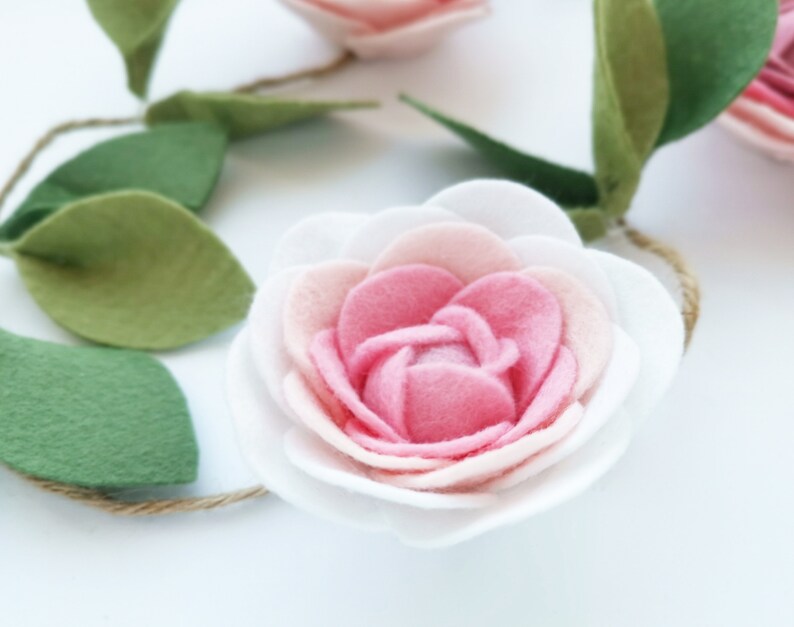 May include: A pink and white felt flower garland with green felt leaves. The flower has multiple layers of petals and is attached to a twine string.