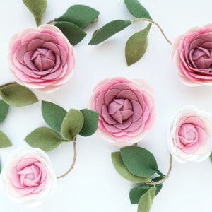 May include: A garland of felt flowers and leaves. The flowers are pink and white and have a layered, ruffled appearance. The leaves are green and have a simple, oval shape.