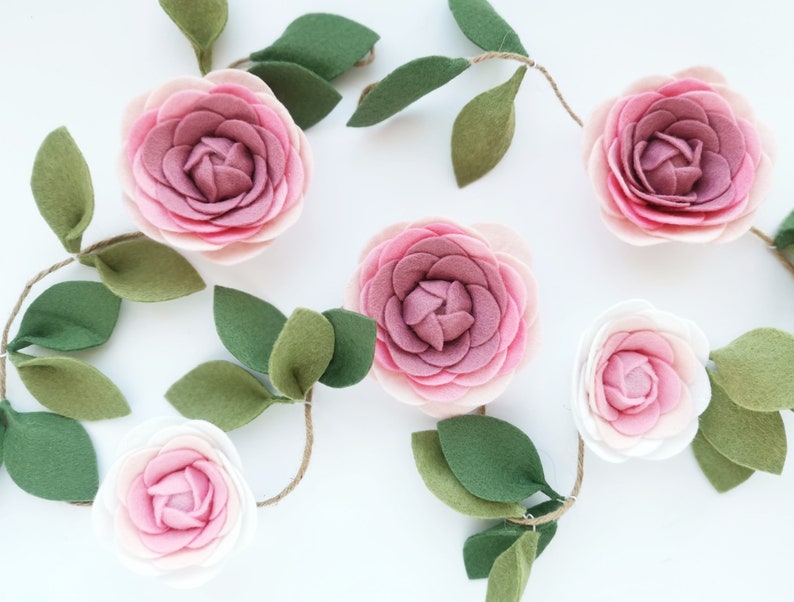 May include: A garland of felt flowers and leaves. The flowers are pink and white and have a layered, ruffled appearance. The leaves are green and have a simple, oval shape.
