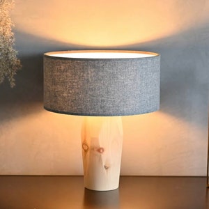 Könnte beinhalten: Eine Tischlampe aus Holz mit einem grauen Stoffschirm. Die Lampe ist eingeschaltet und beleuchtet den Raum. Die Lampe steht auf einem Holztisch mit einer Vase mit getrockneten Blumen.