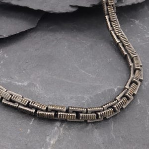 Puede incluir: Un collar de cadena metálica que presenta una serie de eslabones rectangulares entrelazados. Cada eslabón individual tiene una superficie texturizada y estriada con un acabado oscuro y oxidado que resalta el patrón geométrico del metal.