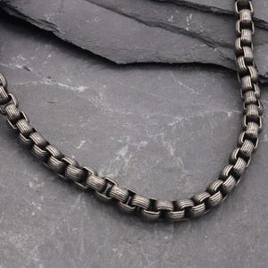 Puede incluir: Un collar de cadena de metal gris oscuro que presenta una serie de eslabones cilíndricos acanalados interconectados. La cadena tiene un acabado mate y oxidado que resalta la superficie texturizada y ranurada de cada eslabón individual.