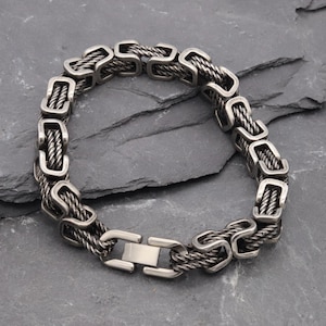 May include: A silver-toned metal bracelet featuring a Byzantine chain design with interlocking rectangular links and a smooth, rectangular clasp closure. The bracelet has a dark, oxidized finish in the recessed areas of the twisted metal links.