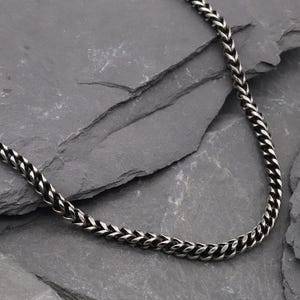 Op de afbeelding: Een zilverkleurige tarweketting bestaande uit een reeks in elkaar grijpende, V-vormige metalen schakels die een getextureerd, gevlochten uiterlijk creëren. De ketting heeft een gepolijste afwerking met donkere accenten in de tussenruimtes.