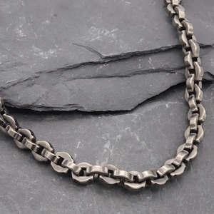 Op de afbeelding: Een ketting in gunmetal grijs met een reeks in elkaar grijpende, afgeronde, dikke schakels. Het metaal heeft een licht verweerde, matte afwerking met zichtbare accenten op de gebogen randen van elke individuele schakel voor een textuur.