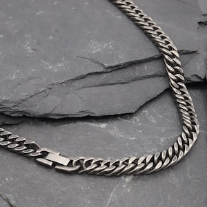 May include: A dark silver-toned curb chain necklace featuring interlocking flat links with a brushed, oxidised finish. The chain includes a rectangular box clasp mechanism that is integrated into the design, creating a continuous look.