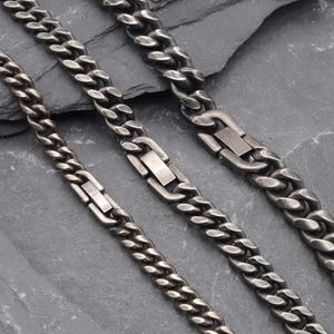 Puede incluir: Tres cadenas de eslabones tipo barbada en gris metalizado oscuro de diferentes anchos, cada una con un cierre rectangular pulido integrado en el diseño. Las cadenas tienen un acabado mate y desgastado, dispuestas en diagonal.
