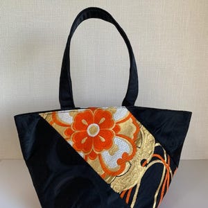 Puede incluir: Una bolsa tote negra con un diseño geométrico con bordado naranja, blanco y dorado. La bolsa tiene dos asas negras.