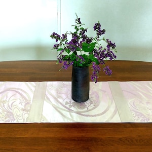 May include: A dark brown vase holds a vibrant arrangement of purple flowers. The vase sits on a light pink and white patterned table runner, placed on a dark wooden table. The scene is set against a neutral background.