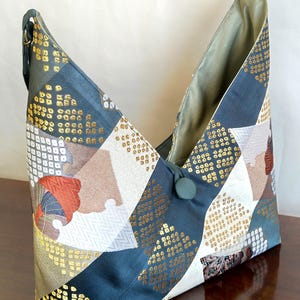 May include: A unique, geometric-shaped tote bag with a patchwork design. The bag features a combination of navy blue, gold, white, and brown fabrics. The interior is a light olive green. A button closure and dark green straps complete the design.