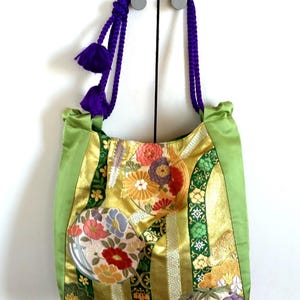 May include: A decorative tote bag with a vibrant floral design. The bag features a gold and green satin fabric with embroidered flowers in shades of red, orange, and blue. It has a purple rope handle with matching tassels.