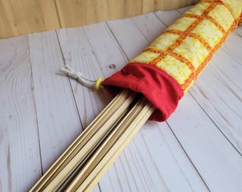 Könnte beinhalten: Eine gelb-rot gemusterte Tasche mit rotem Kordelzug oben, die mehrere hellfarbene Holzlatten enthält. Die Tasche besteht aus Stoff mit einem sich wiederholenden Gitter- und Blumendesign. Der Kordelzug ist weiß mit einem gelben Ring.