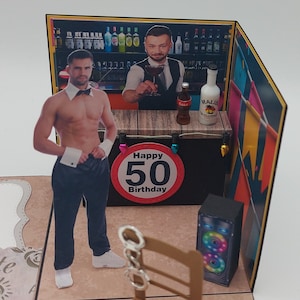 Könnte beinhalten: Eine 3D-Pop-up-Karte mit einem Mann im Smoking und Fliege, der vor einer Bar steht. Die Karte lautet "Happy 50th Birthday" und hat ein rotes und weißes Schild mit der Nummer 50 darauf. Auf der Bar stehen eine Flasche Schnaps und eine Flasche Coca-Cola.