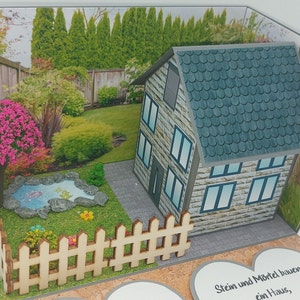 Könnte beinhalten: Eine 3D-Pop-up-Karte mit einem kleinen Haus, einem Zaun, einem Teich und einem Baum. Die Karte enthält eine Botschaft, die lautet "Stein und Mortel bauen ein Haus".
