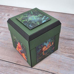 Puede incluir: Una caja verde con ribetes negros e im&aacute;genes de la pel&iacute;cula animada Tarz&aacute;n. La caja tiene una parte superior cuadrada con la palabra "Tarz&aacute;n" impresa. Los lados de la caja presentan im&aacute;genes de personajes y escenas de la pel&iacute;cula.