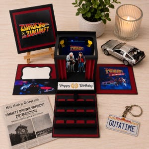 Könnte beinhalten: Eine schwarz-rote Pop-up-Geschenkbox mit Zurück in die Zukunft-Motiven, inklusive Aufstellern von Doc Brown und Marty McFly, einer Hill Valley Telegraph-Zeitung, einem DeLorean-Modellauto und einem California OUTATIME-Nummernschild-Schlüsselanhänger.
