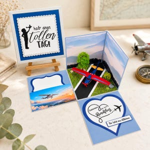 Könnte beinhalten: Eine blau-weiße Pop-up-Grußkarte mit einem Miniatur-Flugzeug in Rot und Blau auf einer Landebahn, einem grünen Baum und einer Bergkulisse. Die Karte enthält den Text "hab' einen tollen TAG!" und "Gutschein für einen Rundflug".