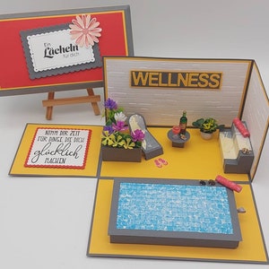 Könnte beinhalten: Eine Pop-up-Karte mit Wellness-Thema. Die Karte ist geöffnet und zeigt einen Miniatur-Pool, Liegestühle und ein Getränk auf einem Tisch. Die Karte hat einen grauen Hintergrund mit gelben Akzenten. Der Text auf der Karte lautet "Wellness" und "Nimm dir Zeit für Dinge die dich glücklich machen".