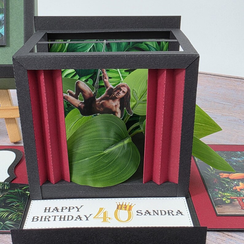 Puede incluir: Una tarjeta negra hecha a mano con tem&aacute;tica de jungla. La tarjeta presenta un recorte de una persona columpi&aacute;ndose en una liana, rodeada de hojas verdes y cortinas rojas. El texto "HAPPY BIRTHDAY 40 SANDRA" se muestra.