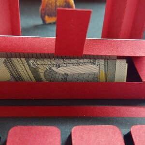 Puede incluir: Una manualidad de papel rojo y negro con una peque&ntilde;a caja rectangular que contiene un billete enrollado. La caja forma parte de una estructura m&aacute;s grande y abierta con detalles rojos y negros. Un anuncio de comida es visible en el fondo.